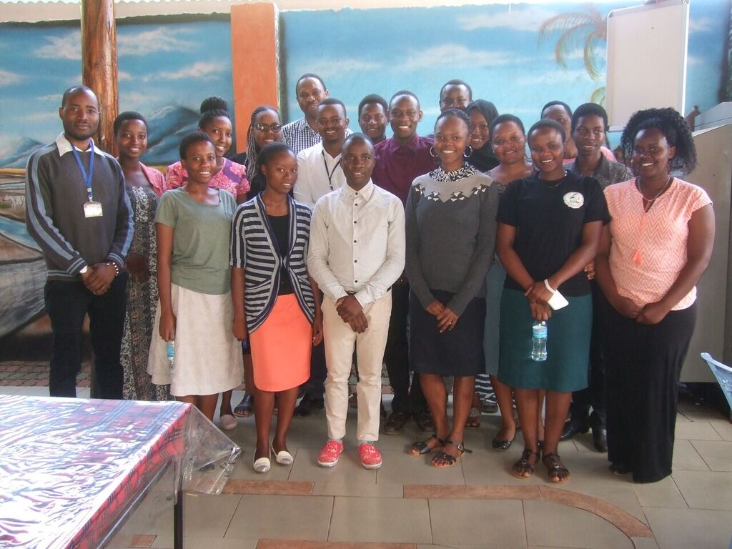 A group of KCMC students and staff standing together for a photo indoors, with a painted mountain mural in the background.